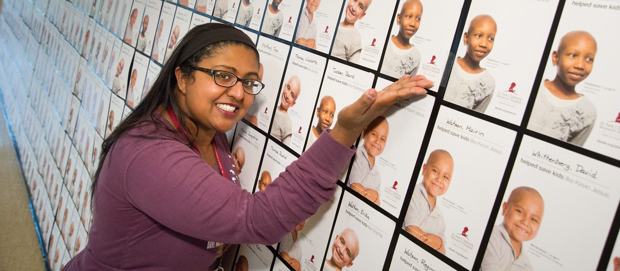 A woman stands next to a wall of St. Jude placards at her workplace and proudly point to one.