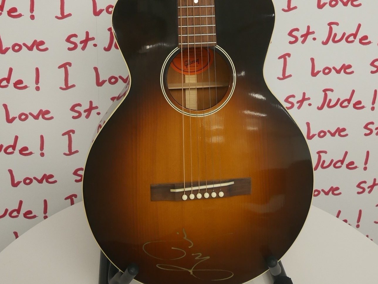 An autographed guitar in front of a background that reads "I love St. Jude"