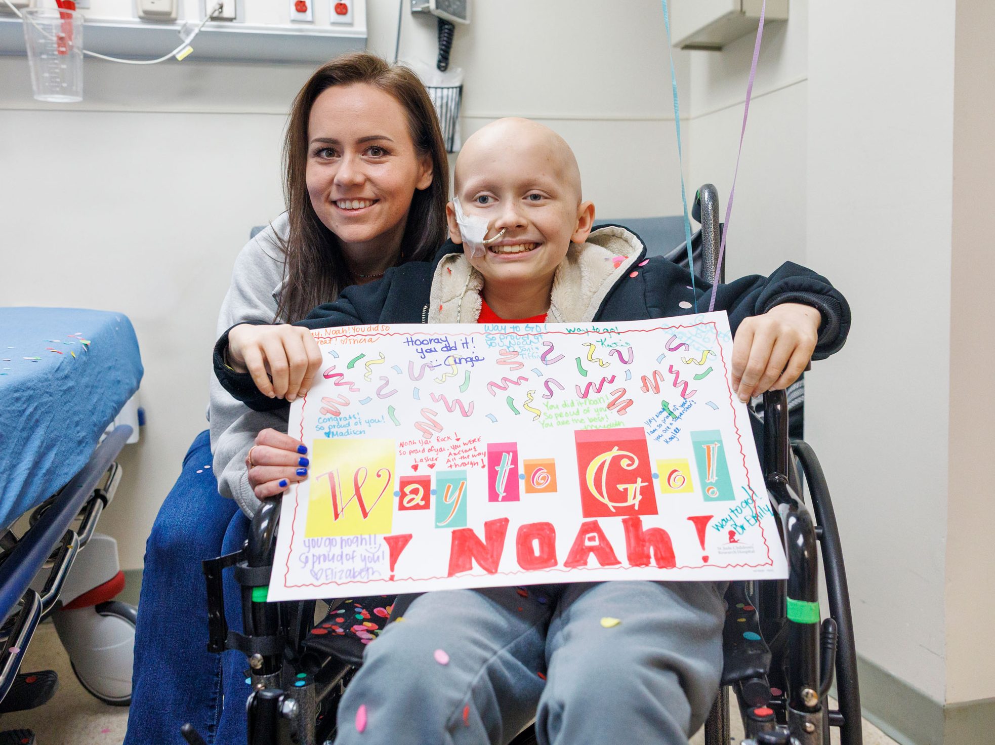 St. Jude patient Noah sits in a wheelchair and holds a sign celebrating the end of chemo while his mother sits behind him.