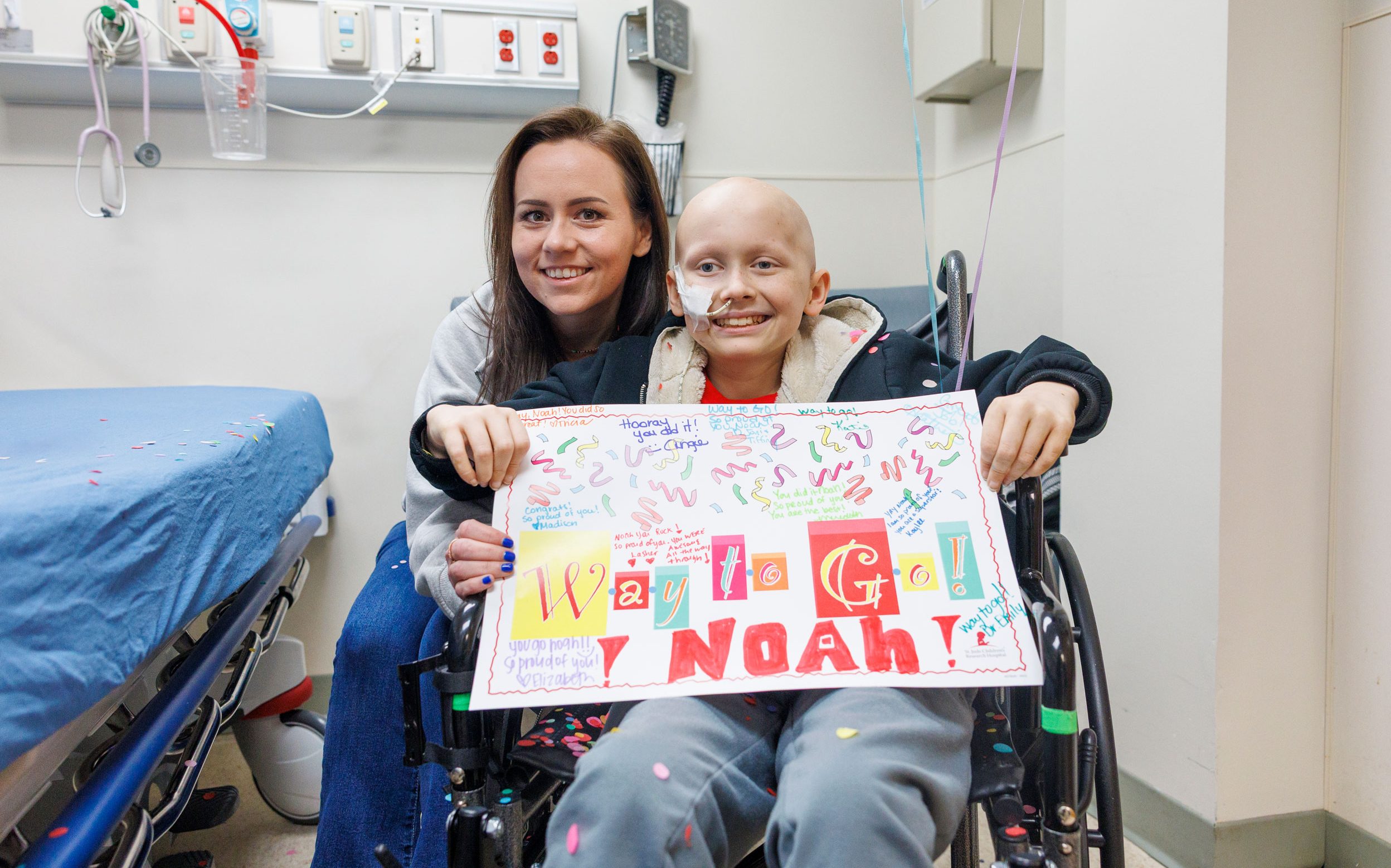 St. Jude patient Noah sits in a wheelchair and holds a sign celebrating the end of chemo while his mother sits behind him.