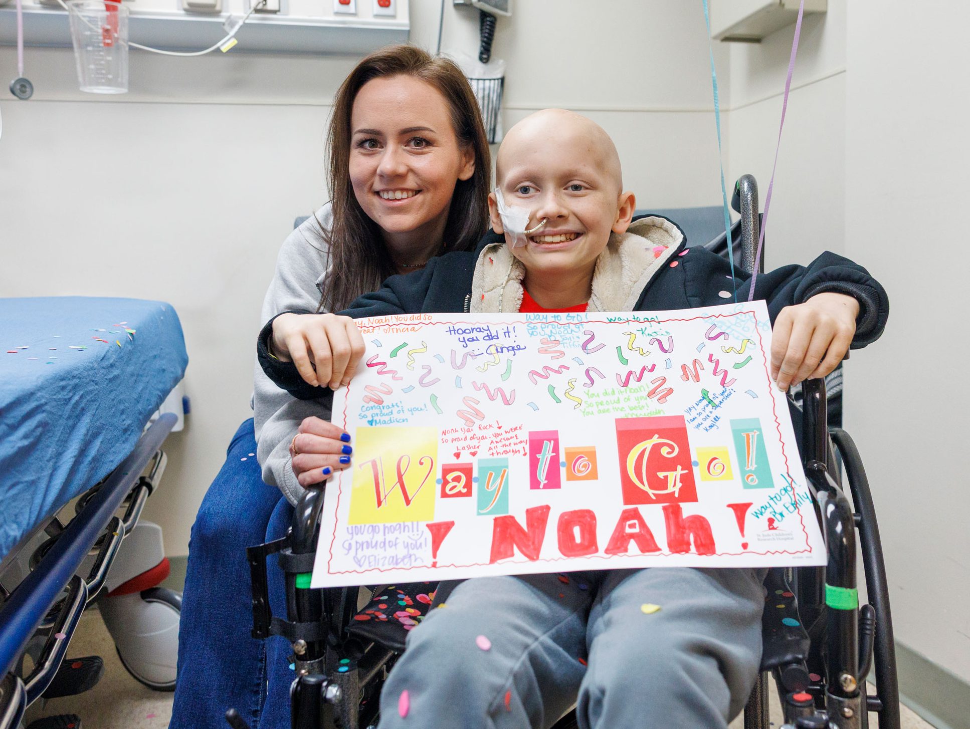 Noah, paciente de St. Jude, celebra el fin de su quimioterapia sosteniendo un letrero mientras está sentado en una silla de ruedas, acompañado por su mamá.
