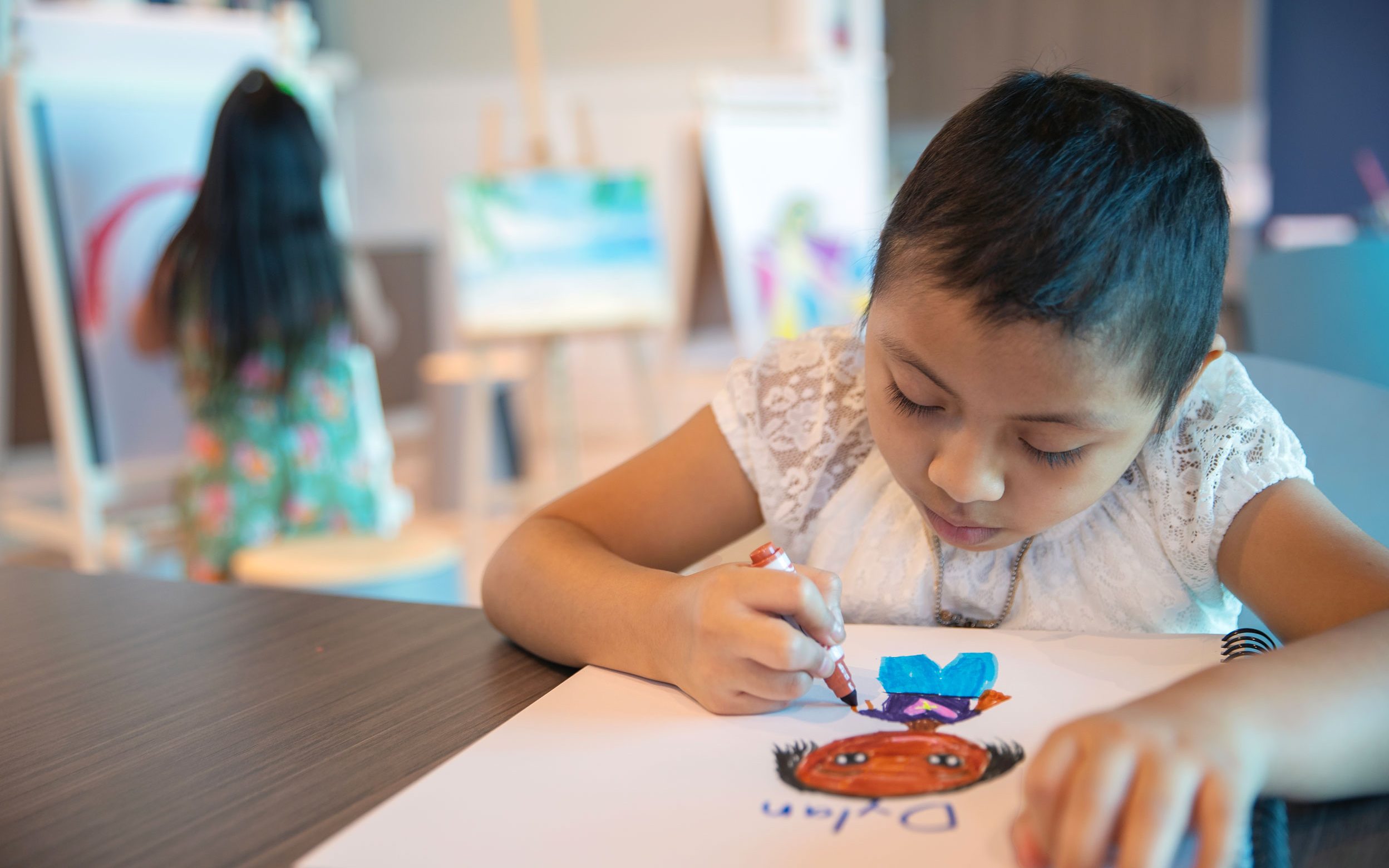 Liliana, paciente de St. Jude, dibujando a una persona, acompañada de su hermana.