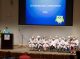 Graduates seated on stage and the School Program director speaking.