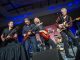 Four men play guitars on stage.