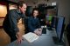 Two male scientists work in front of large microscopes and examine an image