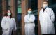 Three researchers in masks and labcoats stand in front of a building.