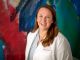 Researcher in lab coat stands in front of a colorful background and smiles.