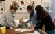 Three nurses in masks practice on mannequin in hospital bed. 