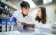 Scientists in white lab coats look at research notebook and discuss findings