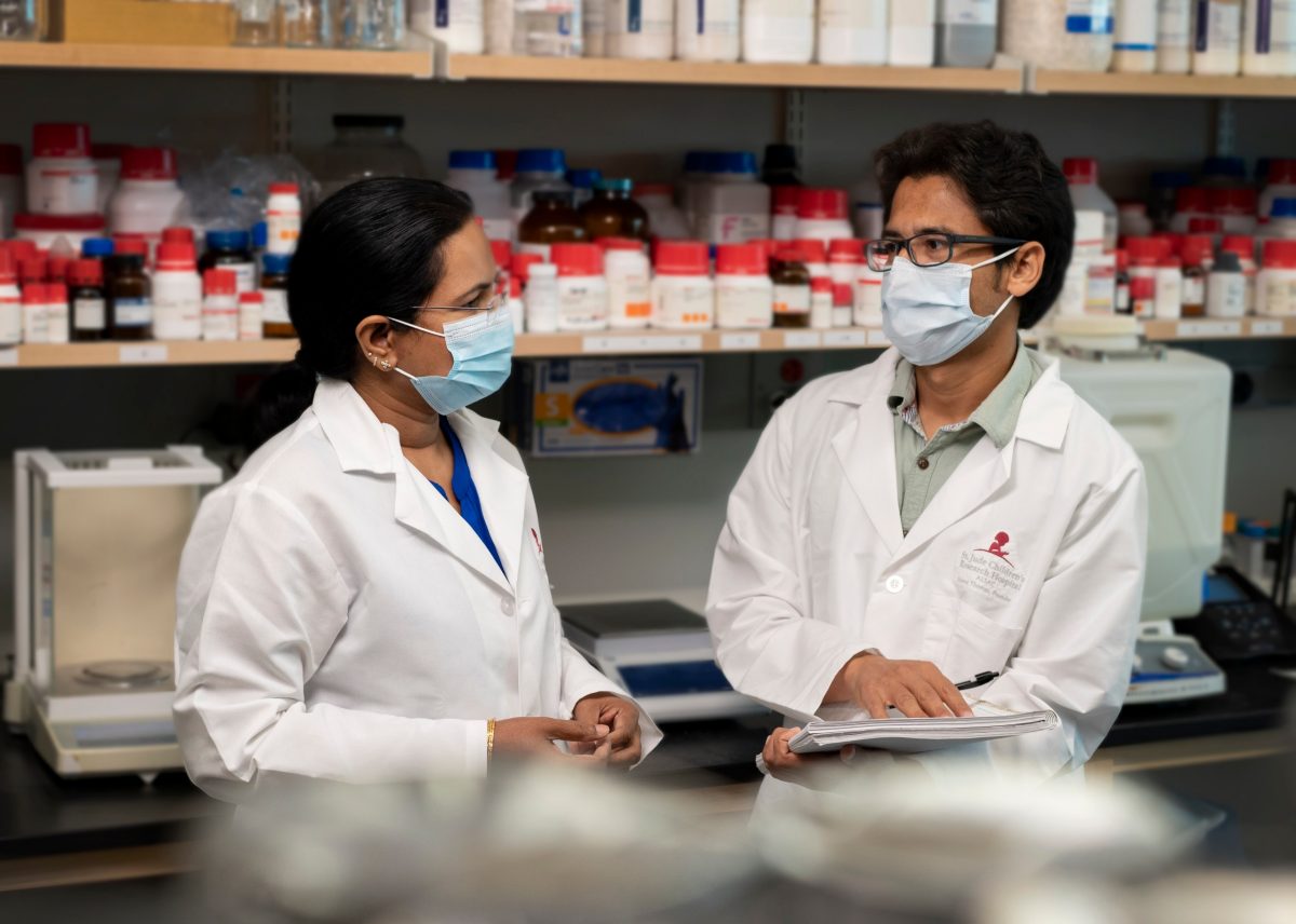 Two scientists wearing lab coats and masks talk in the lab.