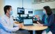 Two researcher in masks are talking while sitting at a desk.
