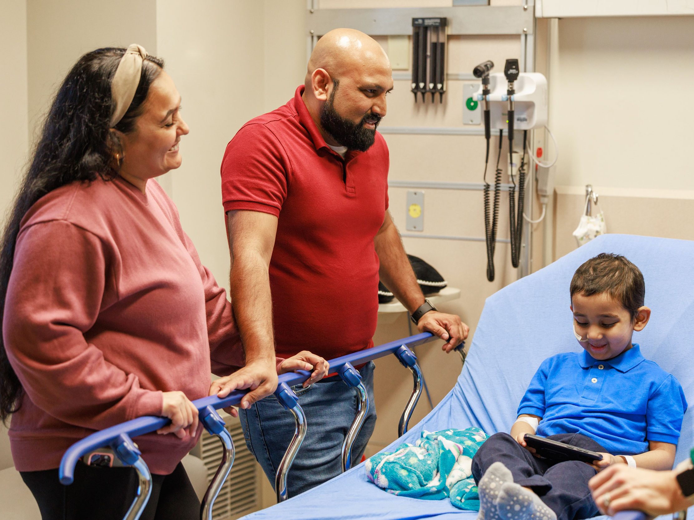 Patient Aditya smiles and plays on a device. The child's parents stand by his bed in a St. Jude treatment room. 